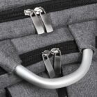 Close-up of gray fabric backpack with multiple zippers and handles. Two pairs of silver zipper pulls are visible, along with a metallic handle in the center. The fabric has a textured pattern, giving it a durable appearance.