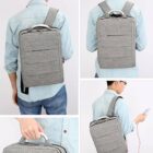 Person wearing a light blue shirt with a light gray backpack. The backpack is rectangular with horizontal stitching and adjustable shoulder straps. The person is seen carrying the backpack, wearing it, and using a smartphone while holding it.