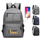 A set of four backpacks in gray and black, with external USB ports. The foreground features a gray backpack with combat impact printed in orange. A smartphone is shown connected to the USB port for charging.