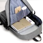 Open gray backpack showing inner contents. Inside, an iPad, laptop, magazine, and small umbrella are visible. There are slots with a smartphone and a white pack in the side pocket. The backpack is on a plain white surface.