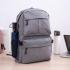 A gray backpack on a desk with multiple compartments and zippers. A blue umbrella is in the side pocket. Nearby are a book, a white thermal flask with a brown lid, and a laptop.