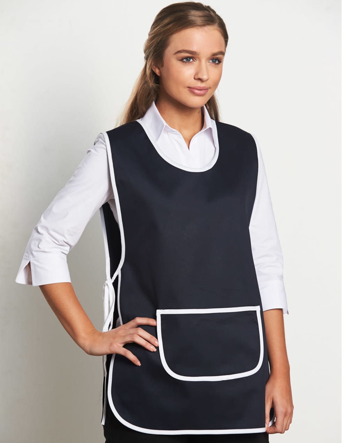A person wearing a black apron with white trim and a large front pocket over a white button-up shirt. The apron is tied at the sides, and the person is standing against a plain white background.