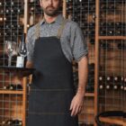 A man wearing a black apron and checkered shirt stands in a wine cellar. He holds a tray with two wine glasses and a bottle. Shelves filled with wine bottles are visible in the background.