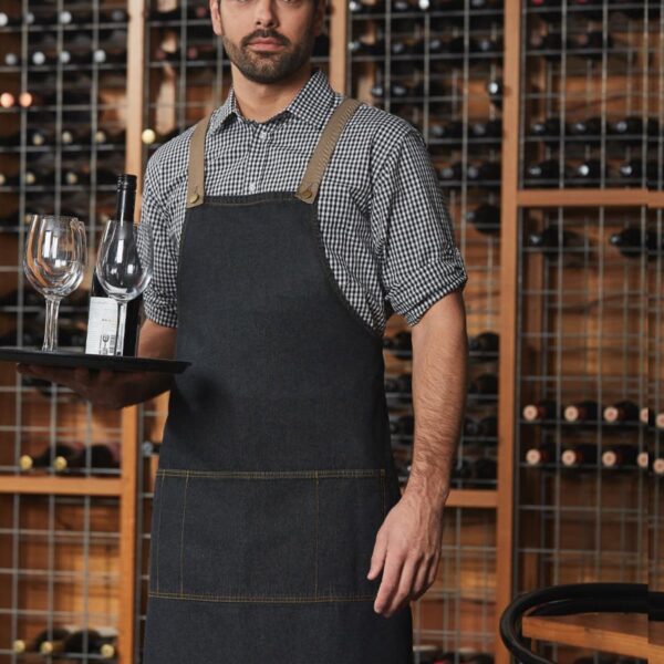 A man wearing a black apron and checkered shirt stands in a wine cellar. He holds a tray with two wine glasses and a bottle. Shelves filled with wine bottles are visible in the background.