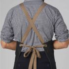 A person is seen from behind wearing a checkered shirt and a dark apron with brown cross-back straps. The apron is tied at the waist with a bow. The background is plain and light gray.