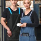 A man and a woman stand side by side in a cafe. Both are wearing black shirts and aprons; the man’s is dark with blue straps, and the woman’s is light denim with striped straps. She holds a card reader. They are smiling softly at the camera.