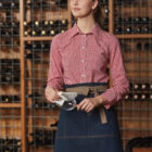 A woman in a red and white checkered shirt with a denim apron stands in front of a wine rack, holding a wine glass. She is looking to the side, with shelves filled with wine bottles in the background.