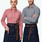 A woman and a man stand side by side, both wearing checkered shirts and dark denim aprons with brown ties. The woman wears a red checkered shirt, while the man wears a blue one. They are smiling at the camera.