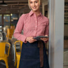 A woman with blonde hair smiles and holds a notepad. She is wearing a red plaid shirt, denim apron, and yellow trousers. She stands inside a cafe with yellow chairs visible in the background.