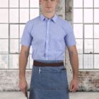 A person wearing a blue checkered shirt and a denim apron stands in front of large windows with a city view. They hold a kitchen knife. The setting appears to be an industrial-style loft or workspace.