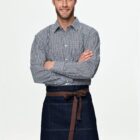 Man wearing a checkered shirt and a denim apron with a brown tie stands smiling with arms crossed against a plain background.