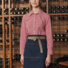 A woman wearing a red checkered shirt and blue apron stands in a wine cellar. Behind her are shelves filled with wine bottles. She is looking forward with a subtle smile.