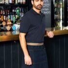 A man stands confidently in front of a bar, wearing a navy polo shirt and a denim apron. He has short dark hair and a beard. Behind him are shelves filled with various bottles.