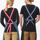 Two people stand side by side, seen from behind. They wear black shirts, khaki pants, and aprons with cross-back straps; one has red and white stripes, the other blue and white. Both have blonde hair tied back.