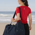A person in a red shirt, black shorts, and a navy cap stands on a beach, facing away. They carry a large navy duffel bag over their shoulder. The ocean and sky are in the background.