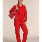 Person smiling, dressed in a red sports outfit including a cap, long-sleeve shirt, and pants. They are holding a matching red and white sports bag over one shoulder and wearing black shoes with white soles, standing against a plain background.