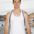 Man in a white tank top standing on a dock, with a blurred waterfront background.