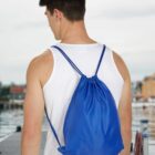 Person in white tank top with blue drawstring bag stands on dock.