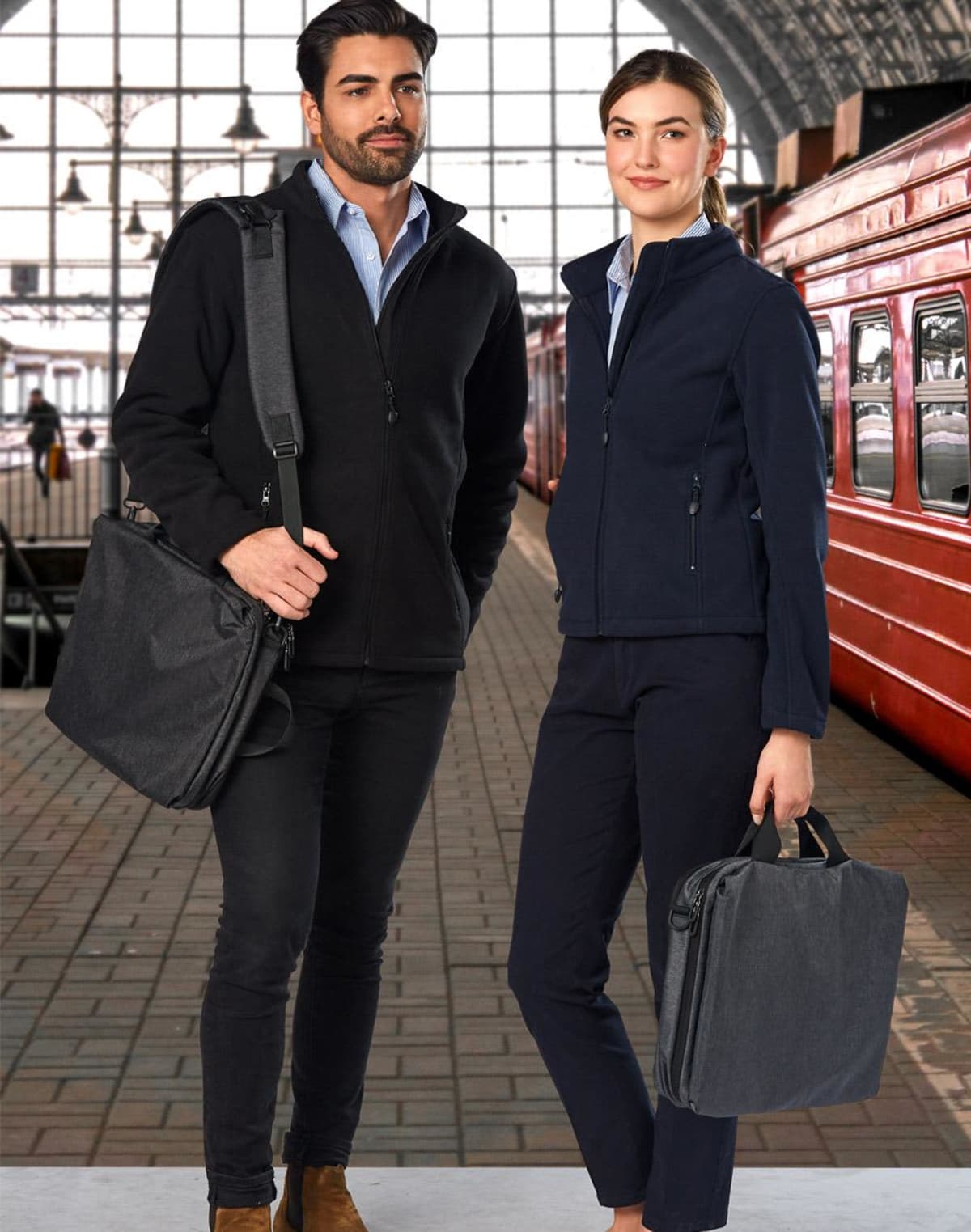 Two people in smart casual outfits with bags standing in a train station.