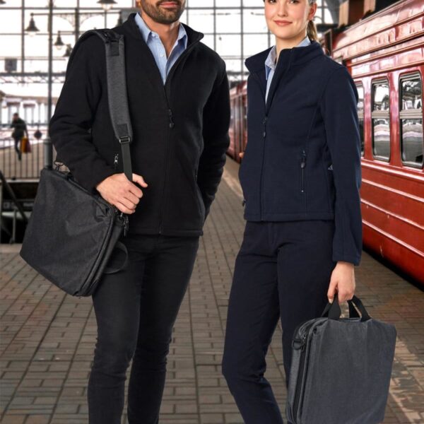 Two people with bags standing on a train platform with trains in the background.