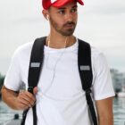 Man with backpack and red cap listens to earbuds outdoors.