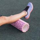 A person using a pink foam roller on their leg while wearing blue running shoes.