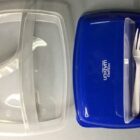 Blue lunchbox with clear compartments and white plastic cutlery on a gray background.