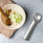 Bowl of green ice cream with pistachios, oats, and a spoon, next to an ice cream scoop.