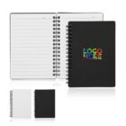 Open black notebook with lined pages and Date header, showing spiral binding. Closed version beside it has a colorful LOGO HERE text on the cover. Additional smaller black and white closed notebooks are also displayed.