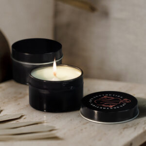Two black tin candles sit elegantly on a table; one is open and lit, casting a warm glow, while the other remains closed. Part of Sheas Favourite autumn merch, these candles bring cozy charm to any setting.