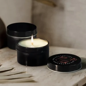 Two black tin candles sit elegantly on a table; one is open and lit, casting a warm glow, while the other remains closed. Part of Sheas Favourite autumn merch, these candles bring cozy charm to any setting.