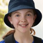 Young girl wearing a navy bucket hat and blue shirt, smiling outdoors.
