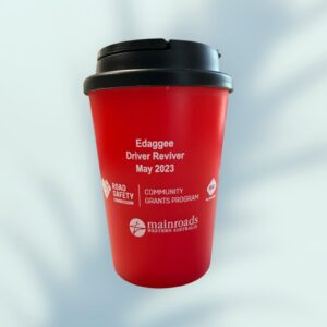 Red reusable coffee cup with a black lid featuring white text: Edaggee Driver Reviver May 2023. Logos include Road Safety Commission, Community Grants Program, and Mainroads Western Australia. Ideal promotional products, these cups blend utility with awareness. The background is a soft gradient.