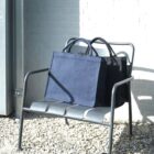 A navy blue tote bag sits on a metal bench in sunlight next to a white brick wall.