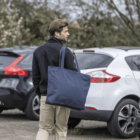 Man carrying a large blue tote bag, walking in a parking lot beside parked cars.