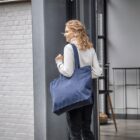 Woman with curly hair carrying a large blue tote bag over her shoulder, walking indoors.