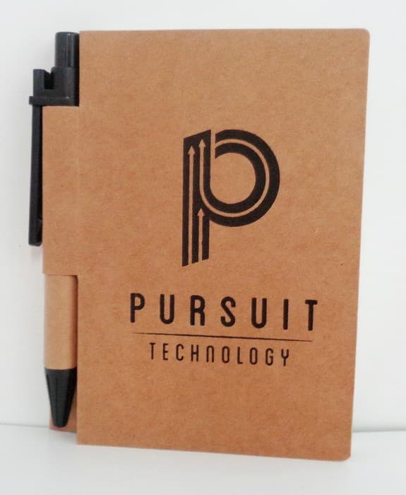 A brown notebook with Pursuit Technology printed on the cover in black. An abstract P logo is above the text. A black pen is attached to the notebooks spine.