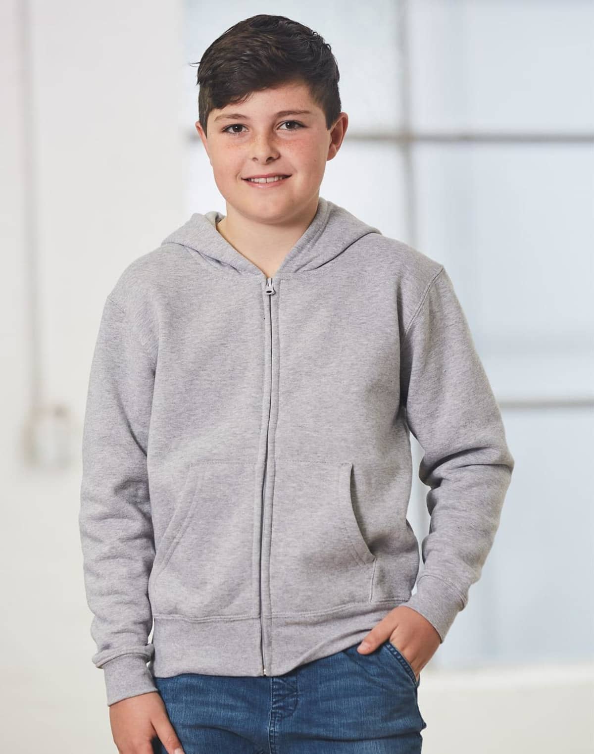 Boy in a gray hoodie and jeans standing indoors, smiling with hands in pockets.