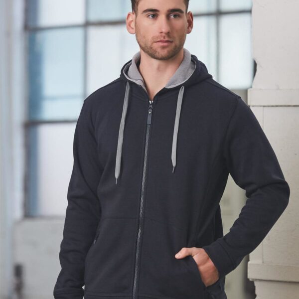 Man in black zip-up hoodie stands by large windows.
