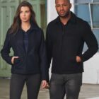 Two people wearing black jackets stand indoors, looking forward.