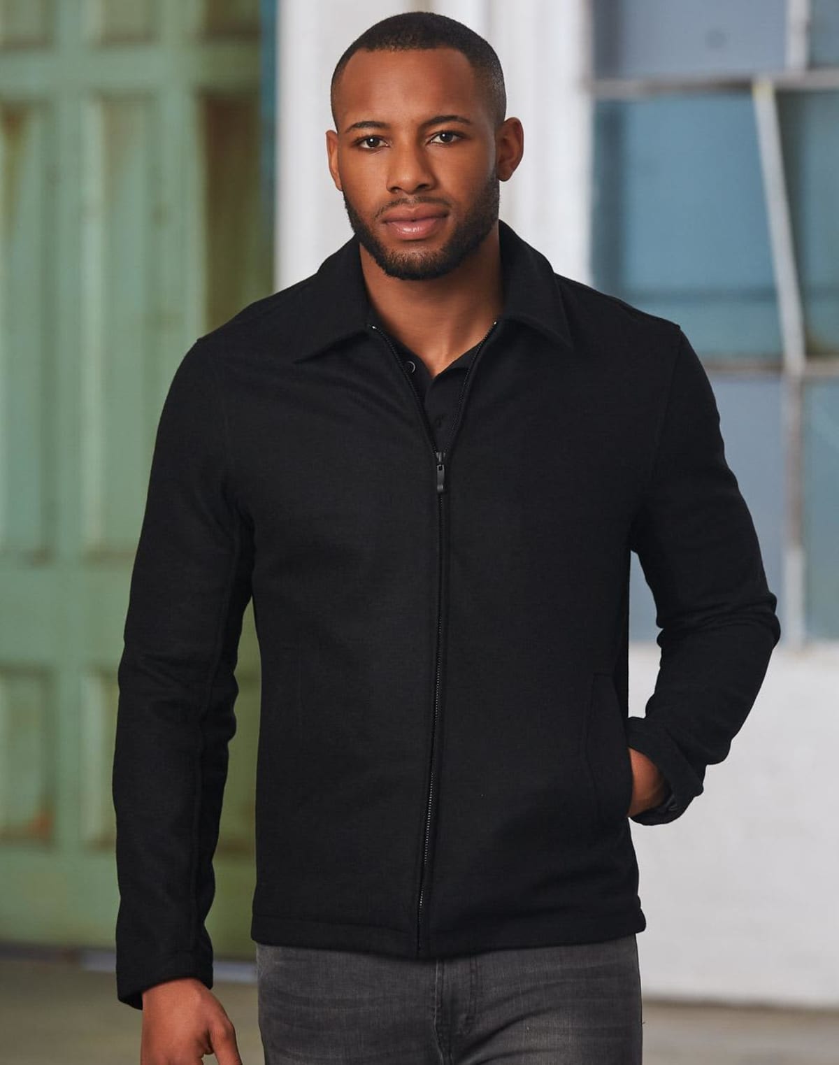 Man in a black jacket and jeans standing indoors with a neutral expression.
