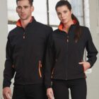 Two people wearing matching black jackets with orange details, standing indoors.