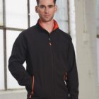 Man in black jacket with orange details standing indoors near large windows.