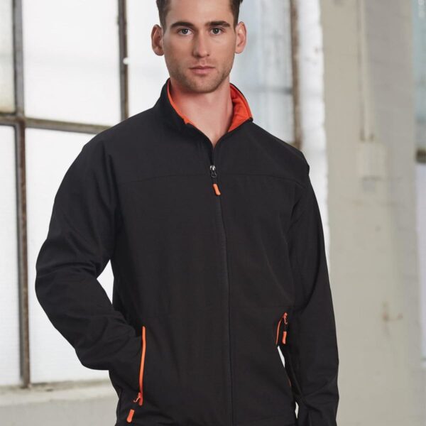 Man in black jacket with orange details standing indoors near large windows.