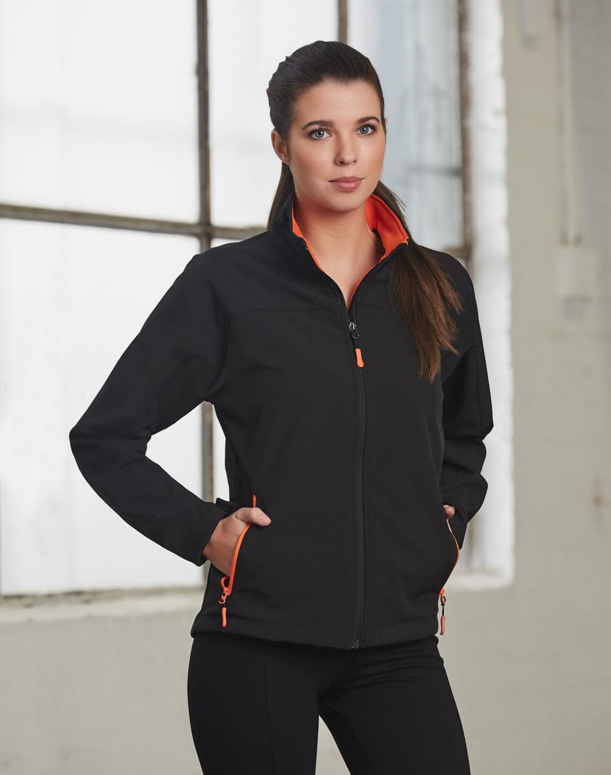 Woman in a black jacket with orange accents stands near a window, hands in pockets.