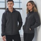 Two people wearing dark gray zip-up jackets, standing by a large window.