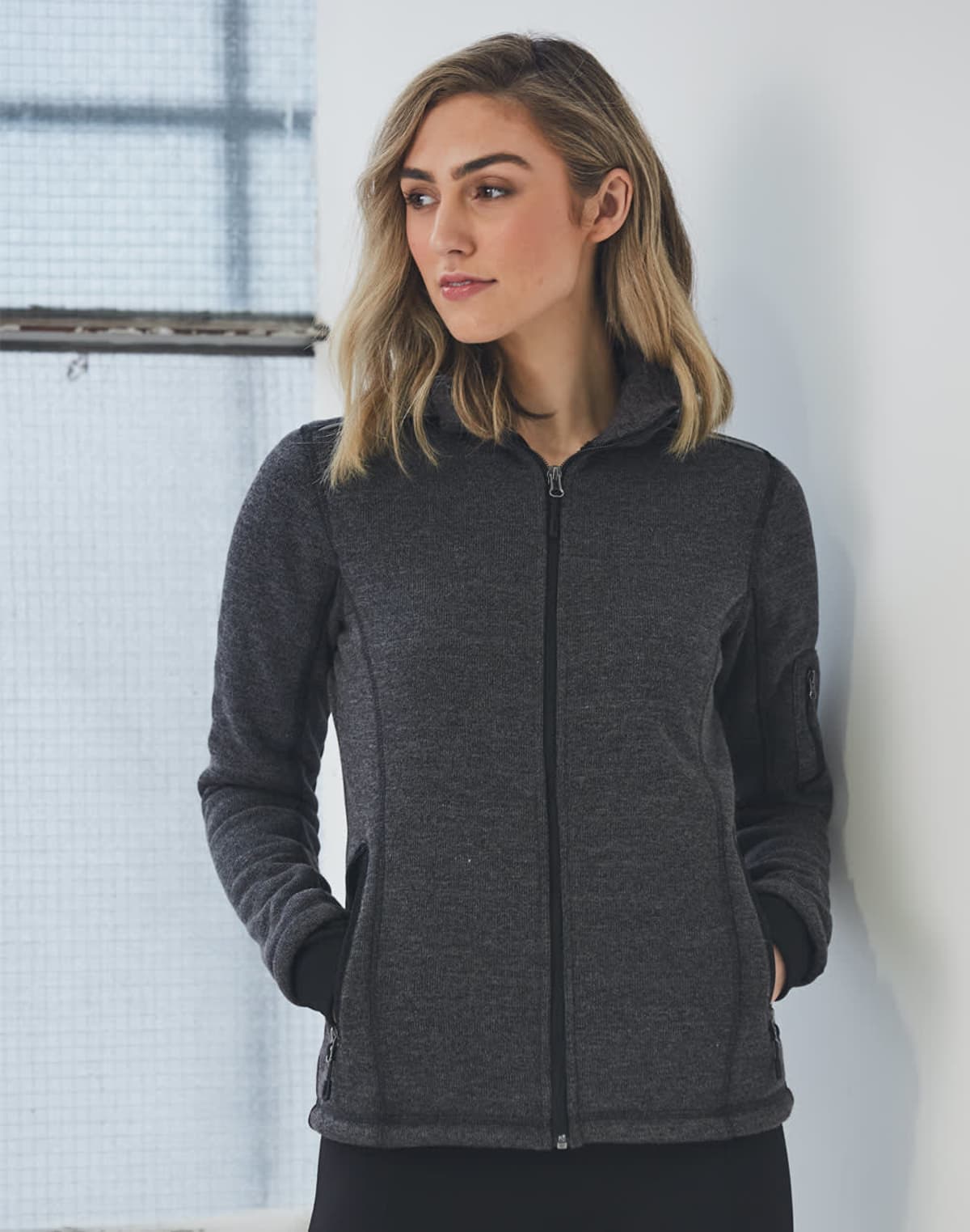 Person in a dark zip-up jacket stands near a window, looking to the side.