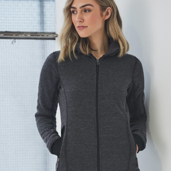 Woman in a gray zip-up jacket standing by a window with hands in pockets.