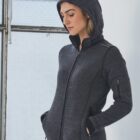 Person wearing a dark gray hoodie, standing by a window with their hands in pockets.