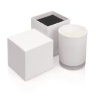 White candle in a glass jar with an open and closed white box next to it on a white background.